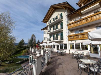 Großes Hotel mit Terrasse, weißen Stühlen, Tischen und Blick auf grüne Wiese und Bäume.