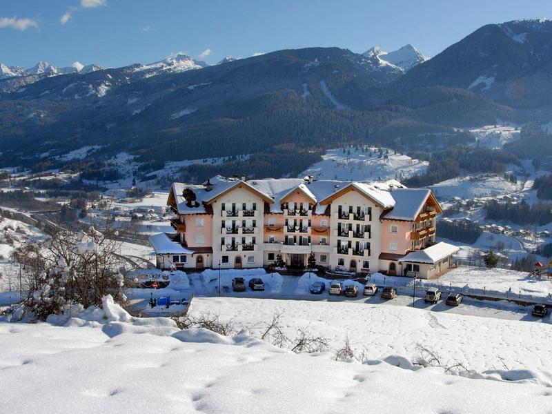 Großes Hotel mit braunen und weißen Fassaden, umgeben von schneebedeckten Bergen und blauem Himmel.
