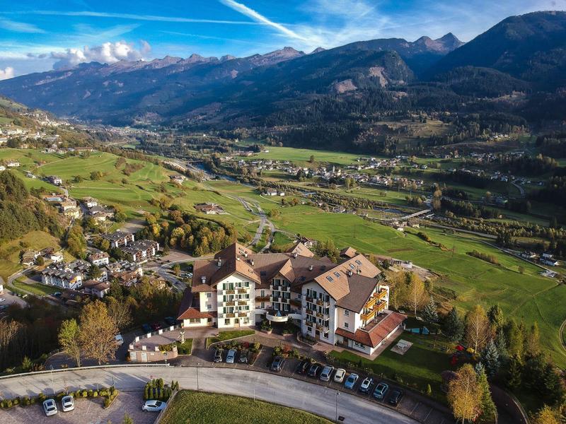 Landschaft mit grünen Feldern, Bergen im Hintergrund und einem Gebäude mit umliegenden Parkplätzen.