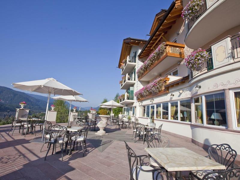 Terrassenrestaurant mit Tischen, Stühlen, Sonnenschirmen und Blumen an einem mehrstöckigen Gebäude.