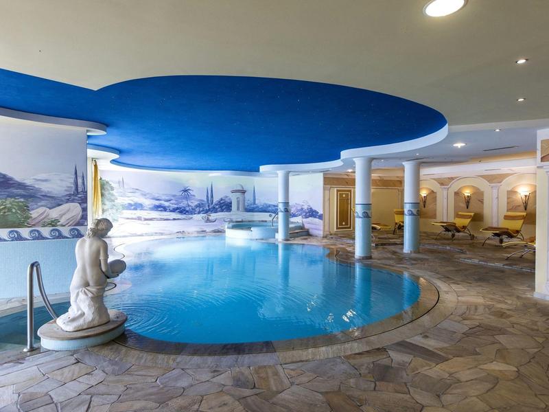 Innenraum mit rundem Schwimmbecken, blauer Decke, Statuen und Mosaiksteinboden.