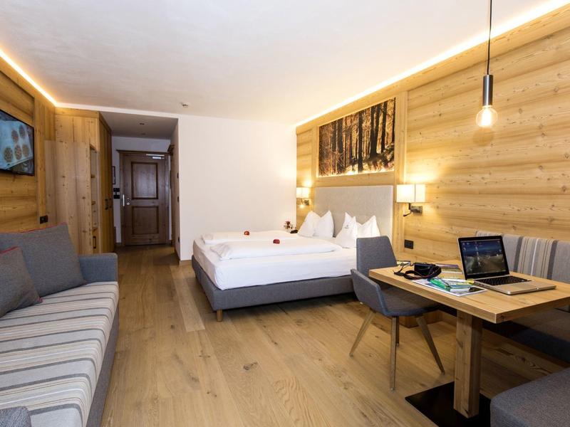 Hell eingerichtetes Hotelzimmer mit Holzwänden, Doppelbett, Sofa und Esstisch mit Laptop.