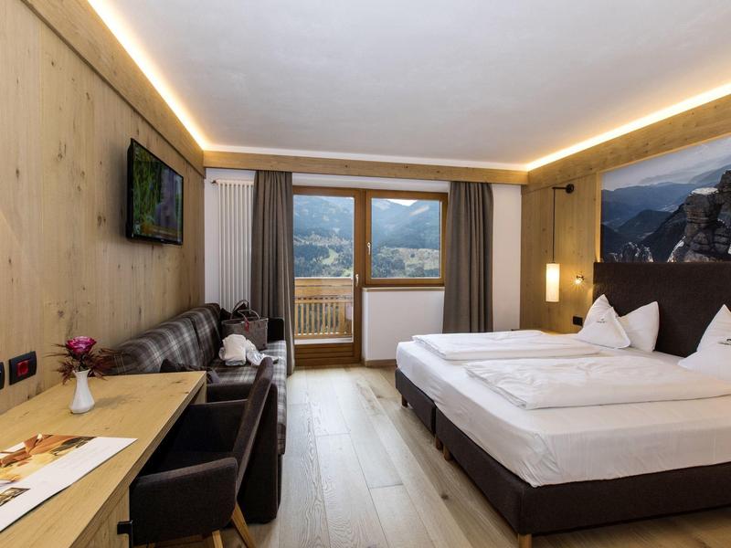 Modernes Hotelzimmer mit Doppelbett, Sofa, Schreibtisch und Balkon, helle Holzböden und warme Beleuchtung.