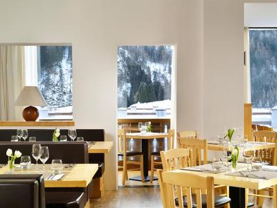 Modernes Restaurant mit Holztischen, Stühlen und großen Fenstern, die Berglandschaft zeigen.