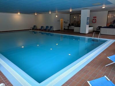 Innen-Schwimmbad mit blauem Wasser, rutschfestem Rand und mehreren Liegestühlen entlang der Wand.