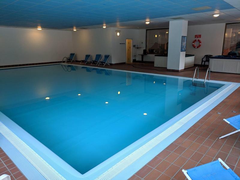 Innen-Schwimmbad mit blauem Wasser, rutschfestem Rand und mehreren Liegestühlen entlang der Wand.