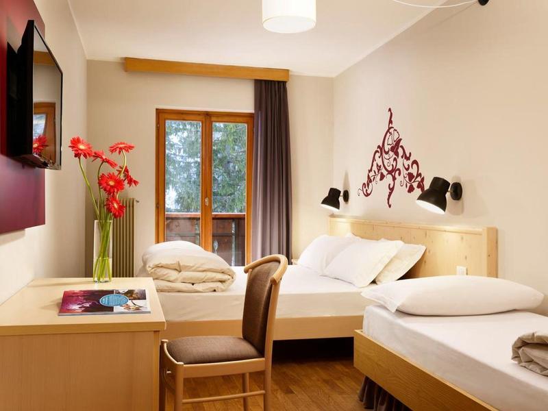 Helles Hotelzimmer mit zwei Einzelbetten, Holzmöbeln, großer Vase mit roten Blumen und Balkonfenster.