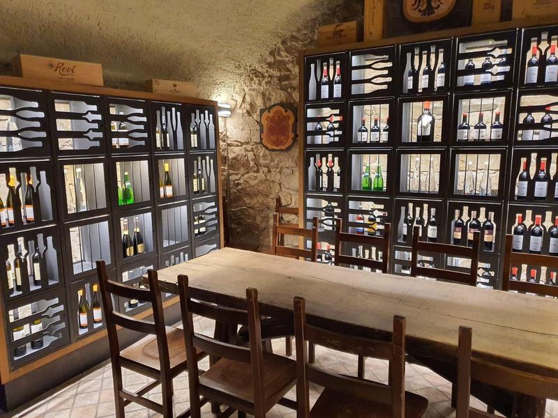 Kellerraum mit Holzregalen voller Weinflaschen und einem langen Holztisch mit Stühlen in der Mitte.