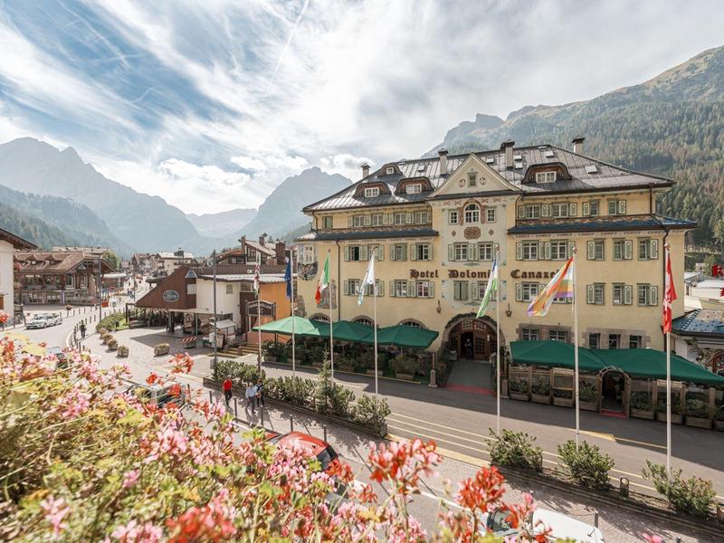 Bergdorf mit pastelgelben Gebäuden, grünen Markisen, blühenden Blumen und Bergen im Hintergrund.