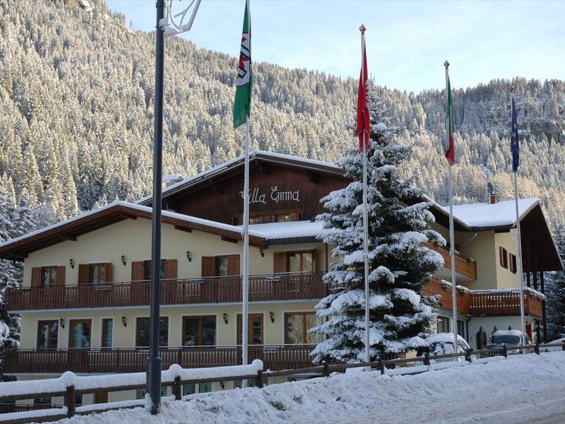 Verschneites Holzhaus mit drei Fahnen und Tannenbaum vor bewaldeten, schneebedeckten Bergen.