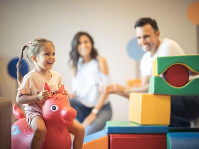 Kleines Mädchen spielt auf einem roten Gummitier, Eltern beobachten im Hintergrund.