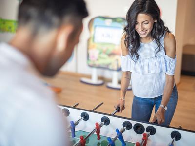 Zwei Erwachsene spielen Tischkicker, Frau lächelt, Mann ist unscharf im Vordergrund, heller Raum.