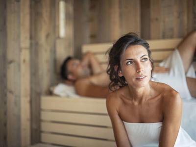Frau in weißem Handtuch sitzt vor Holzsauna, Mann liegt entspannt dahinter auf Holzbank.