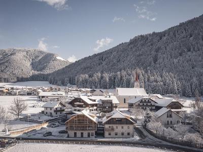 Verschneites Dorf mit Häusern, Bergen und Wald im Hintergrund unter blauem Himmel.