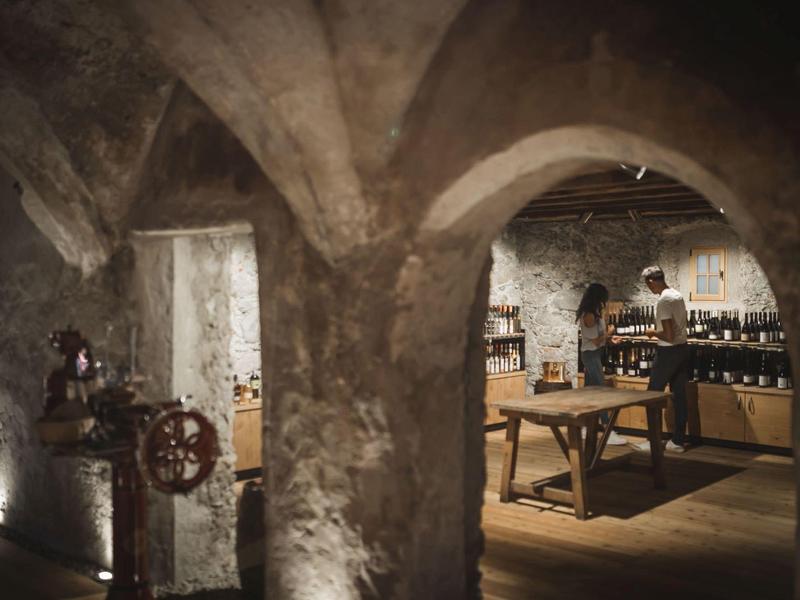 Alte Kellergewölbe mit Steinsäulen und Holztischen, auf denen Weinflaschen stehen.