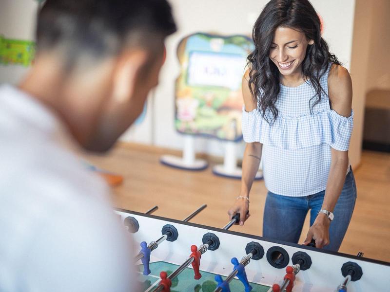 Zwei Erwachsene spielen Tischkicker, Frau lächelt, Mann ist unscharf im Vordergrund, heller Raum.