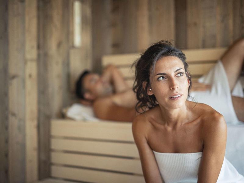 Frau in weißem Handtuch sitzt vor Holzsauna, Mann liegt entspannt dahinter auf Holzbank.