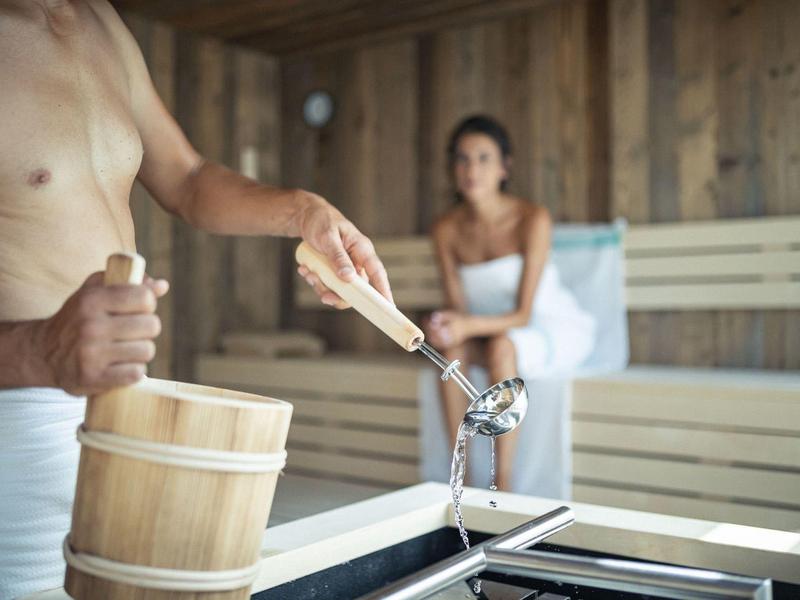 Mann hält Kelle und Eimer, Frau sitzt auf Holzbank in rustikaler Sauna.
