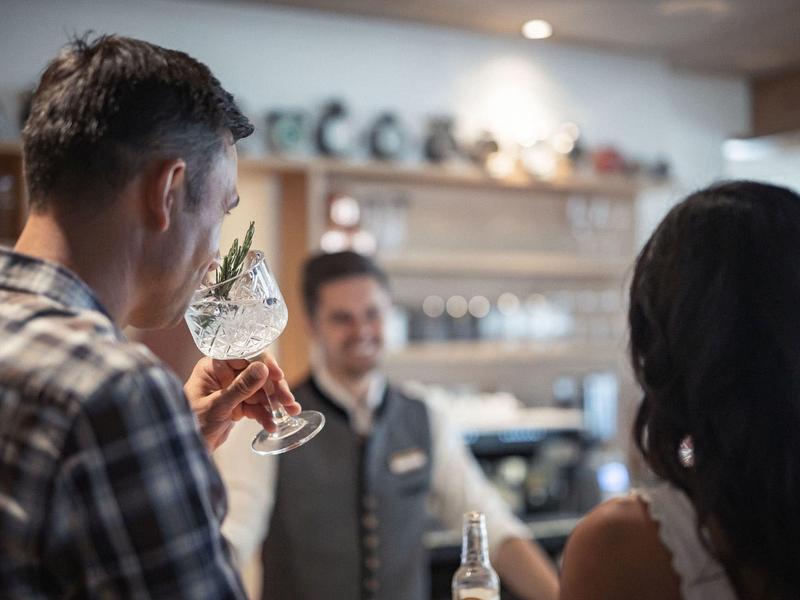 Zwei Personen an einer Bar, die ein Getränk probieren, während ein Barkeeper lächelnd im Hintergrund steht.
