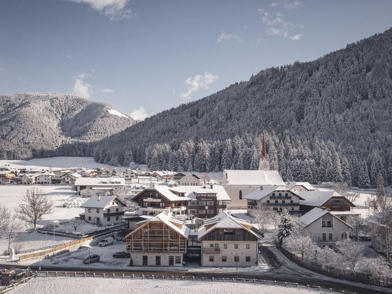 Verschneites Dorf mit Häusern, Bergen und Wald im Hintergrund unter blauem Himmel.