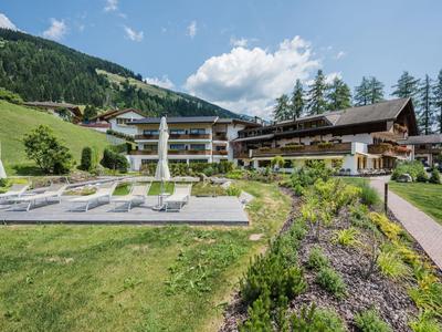 Großes Berghotel mit Sonnenterrasse, Liegestühlen und grüner Wiese, umgeben von Bäumen und Bergen.