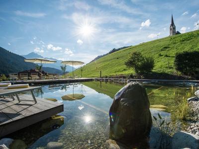 Ein sonniger Bergsee mit steinigem Ufer, Liegestühlen und einer entfernten Kirche auf dem Gras.