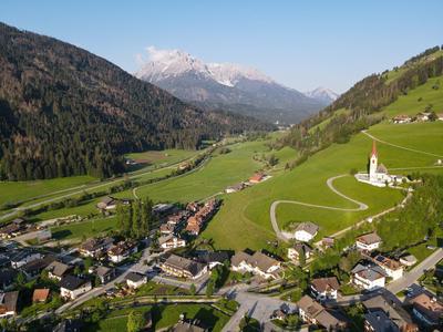 Grünes Tal mit Dorf, Kirche auf Hügel, gewundene Straße und schneebedeckte Berge im Hintergrund.