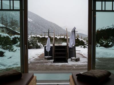 Blick aus Innenraum auf verschneite Terrasse mit Holztreppe, Tüchern und Bergen im Hintergrund.