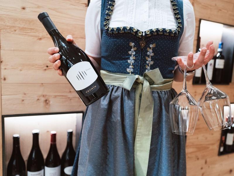 Frau im Dirndl hält Weinflasche und zwei Gläser in einem holzvertäfelten Raum mit Weinregalen.