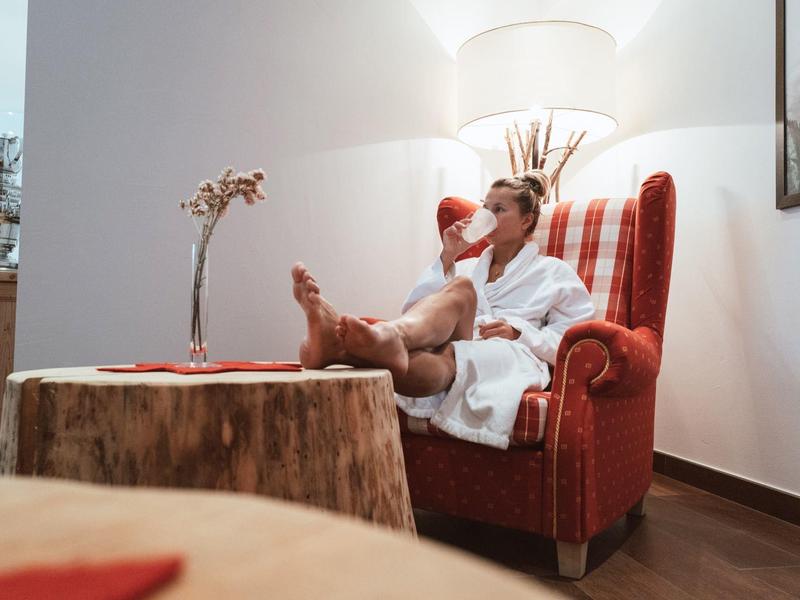 Ein Mann im weißen Bademantel sitzt entspannt in einem roten Ohrensessel vor einem Holzscheiben-Tisch.