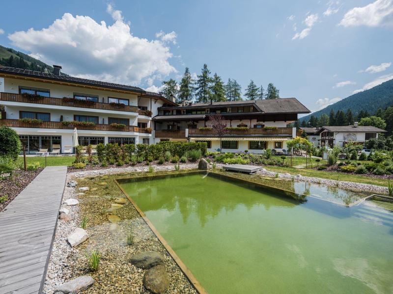 Hotelgebäude neben grünlich klarem Naturteich mit Steg und Bergen im Hintergrund bei blauem Himmel.