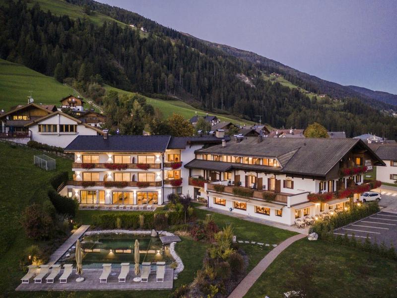 Ein beleuchtetes Berghotel mit Garten, Pool und umliegenden grünen Hügeln bei Abenddämmerung.