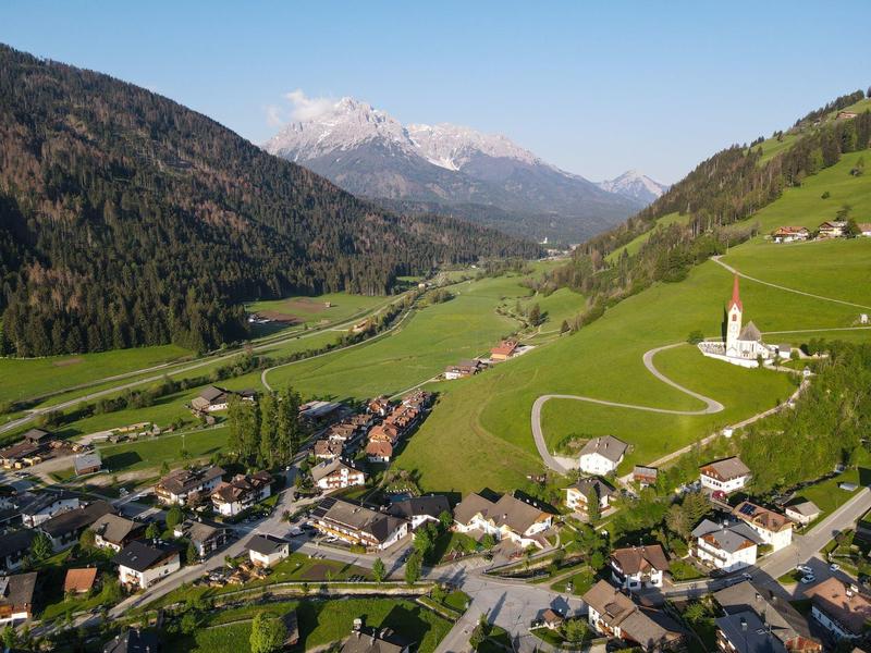 Grünes Tal mit Dorf, Kirche auf Hügel, gewundene Straße und schneebedeckte Berge im Hintergrund.