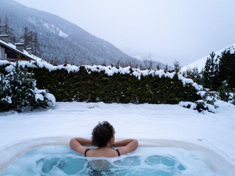 Person entspannt im warmen Whirlpool, umgeben von schneebedecktem Garten und Bergen im Hintergrund.