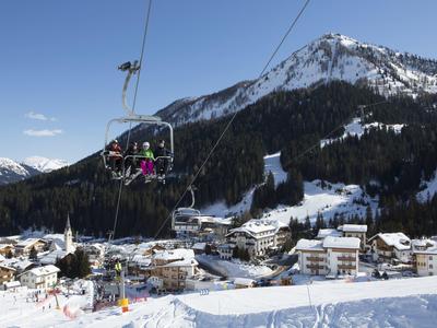 Skifahrer auf Sessellift über schneebedecktem Bergdorf und Bergen bei klarem Himmel.
