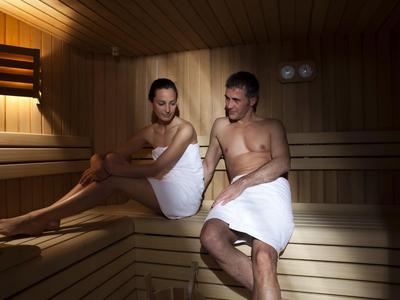 Ein Mann und eine Frau sitzen in einem Saunaraum aus hellem Holz, beide mit weißen Handtüchern bedeckt.