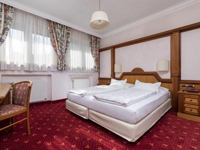 Einzelzimmer mit Doppelbett, rotem Teppich, Holzmöbeln und floral gemusterten Vorhängen.