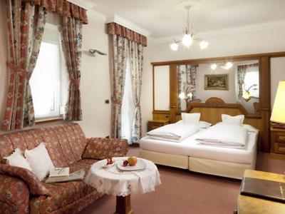 Wohnzimmer mit braunem Sofa, rundem Tisch und angrenzendem Schlafzimmer mit Doppelbett.