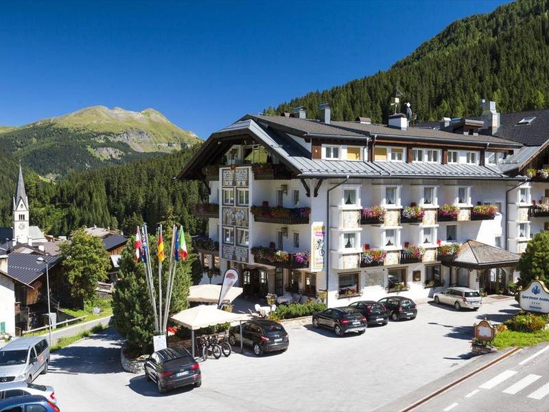 Großes Hotel mit Balkonen, Autos davor, Berge und Wald im Hintergrund, blauer Himmel.