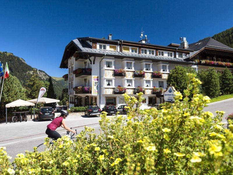 Großes Hotelgebäude mit Balkonen, umgeben von Bergen, blauem Himmel und gelben Blumen im Vordergrund.