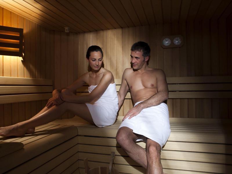 Ein Mann und eine Frau sitzen in einem Saunaraum aus hellem Holz, beide mit weißen Handtüchern bedeckt.