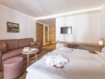 Gemütliches Hotelzimmer mit Bett, Sofa, Stehlampe, TV und Holzmöbeln in warmen Farben.