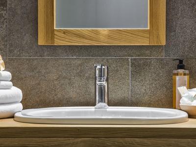 Weißes Waschbecken mit Chromwasserhahn, Handtücher links, Seifenspender und Schalen rechts auf Holzplatte.