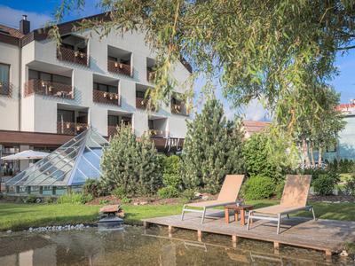 Hotelgarten mit Teich, Holzdeck und zwei Liegestühlen, umgeben von Bäumen und Gebäuden.