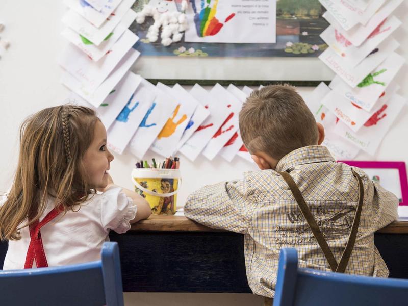 Zwei Kinder sitzen nebeneinander an einem Tisch, umgeben von bunten Papierfahnen und Bildern an der Wand.