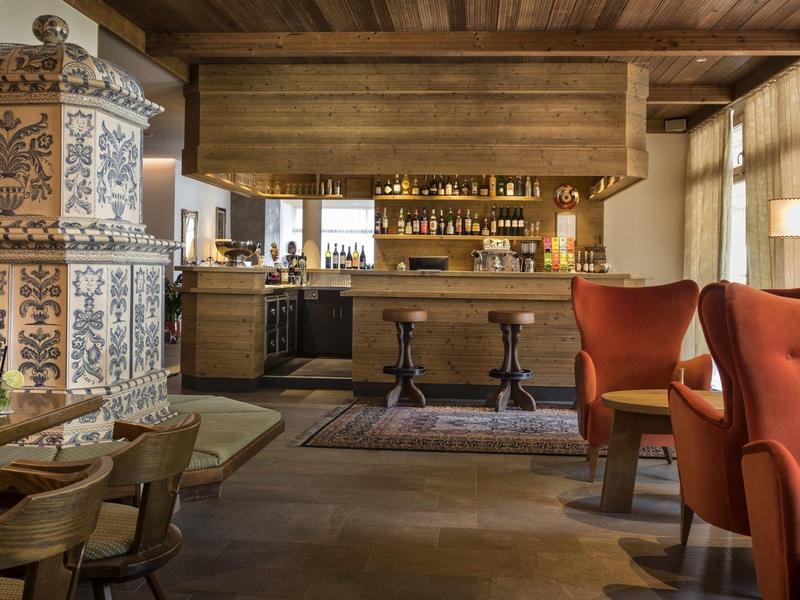 Gemütliche Gaststube mit Holzbalken, buntem Kachelofen, roten Sesseln und Holztischen.