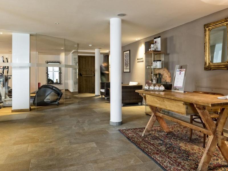 Großer Raum mit Steinfussboden, Holztisch, Teppich, Säule und Fitnessgeräten im Hintergrund.