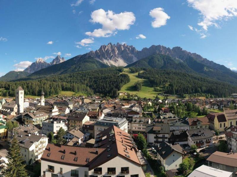Panorama einer sonnigen Stadt mit roten Dächern, Bergen im Hintergrund und blauem Himmel mit Wolken.