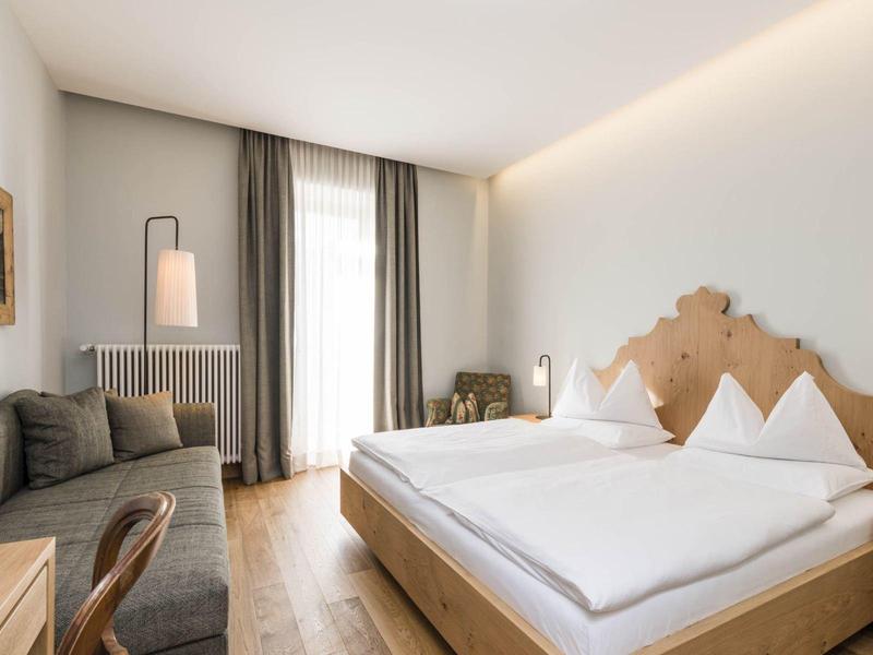 Helles Schlafzimmer mit großem Bett, grauem Sofa, Holzfußboden und hohen Fenstern mit Gardinen.