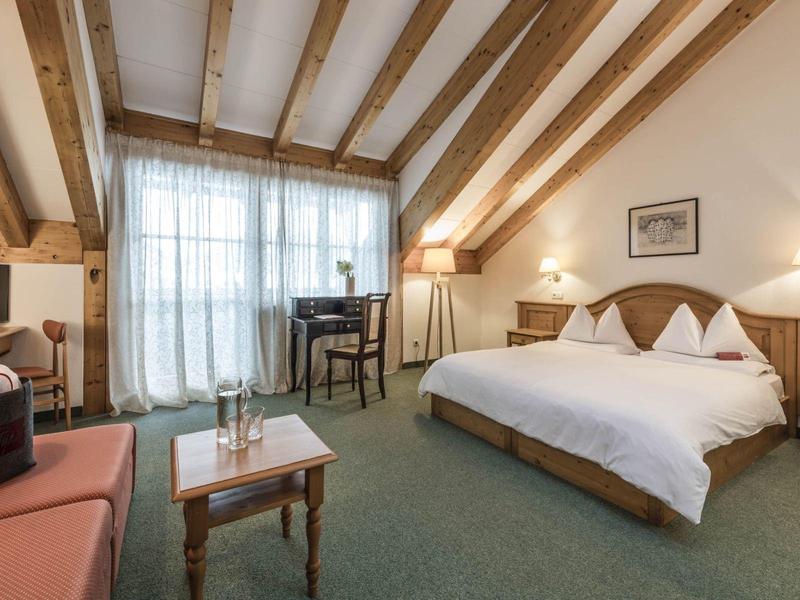 Gemütliches Hotelzimmer mit Holzbalken, großem Bett, Tisch, Stühlen und Balkonfenstern.